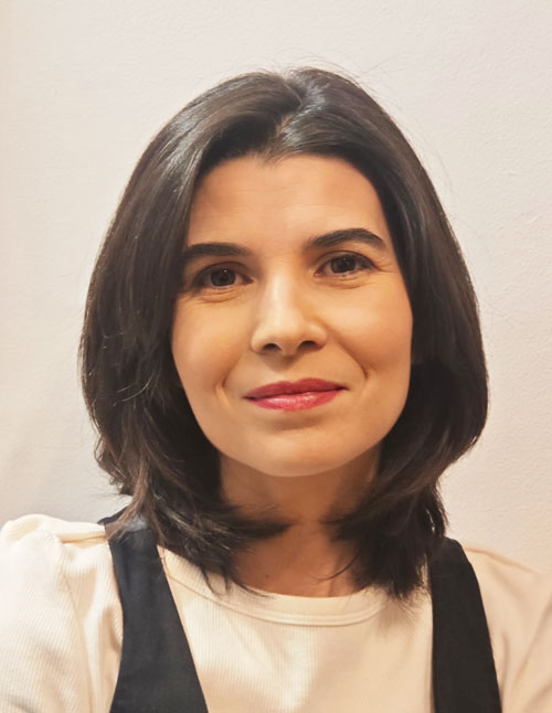 Una mujer con el pelo oscuro hasta los hombros, vestida con una camiseta blanca y un chaleco negro, sonríe con dulzura frente a un fondo liso y claro, lo que evoca el ambiente acogedor de una sala de integración sensorial en Madrid.